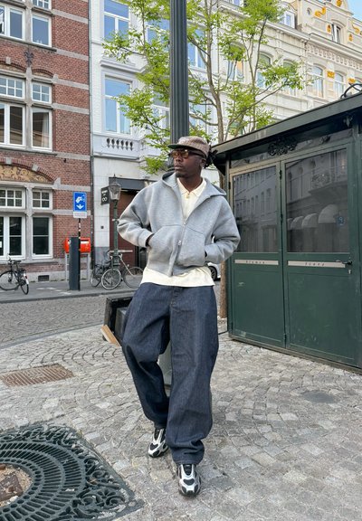 Sudadera gris con cremallera, camiseta blanca, vaqueros oscuros holgados, gorra con estampado y calzado distintivo, con un fondo urbano.