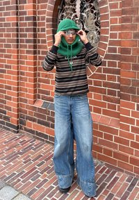 Zelena pletena kapa s vezicom, crna prugasta majica s dugim rukavima i široke plave traperice. Crne čizme na popločanoj stazi.