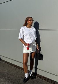 T-shirt bianco oversize, mini gonna bianca, mocassini neri, calze bianche, borsa nera, che tiene in mano una lattina di bevanda rossa, in piedi contro un muro grigio.
