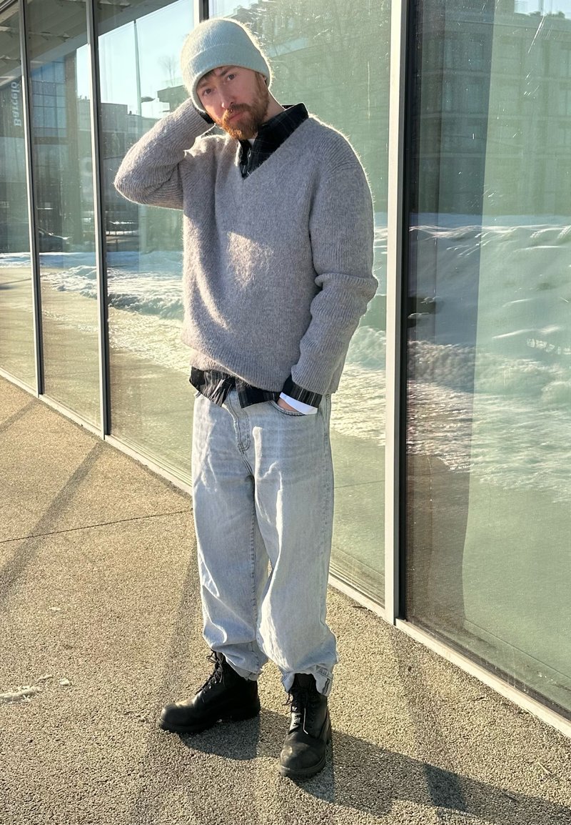 Mann mit grauer Mütze, grauem Pullover, hellen Jeans und schwarzen Stiefeln steht draußen in der Sonne an einer Glaswand, eine Hand am Kopf, die andere in der Tasche.