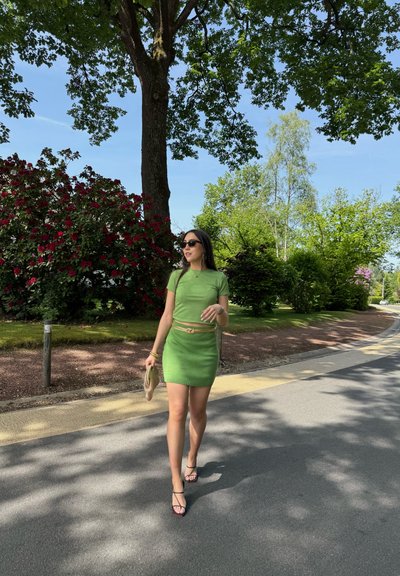 Mujer con vestido verde y gafas de sol caminando por un camino sombreado bordeado de árboles y arbustos floridos en un día soleado.