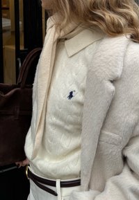 Pull crème en maille torsadée avec un petit logo, superposé sous un manteau beige clair, accompagné d'une écharpe douce et d'un sac marron foncé.