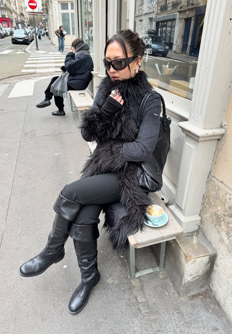 Femeie cu cizme negre, ochelari de soare și fulard de blană stând pe o bancă în fața unei cafenele, cu o ceașcă de latte alături.