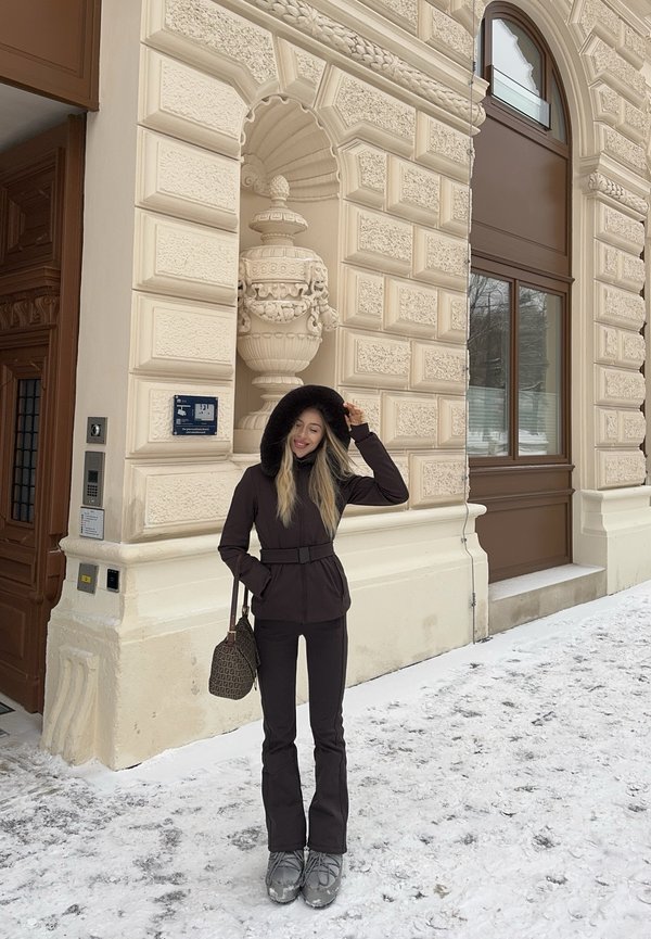 Žena v čiernom zimnom kabáte s kapucňou z kožušiny, čiernych nohaviciach, sivých čižmách a s kabelkou s potlačou stojí na zasneženej ulici pri zdobenej béžovej budove.