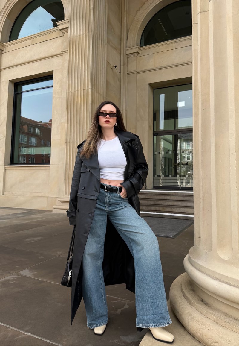 Femme portant un trench-coat en cuir noir, un crop top blanc, un jean à jambes larges, des bottes blanches et des lunettes de soleil, debout près d'une colonne en pierre à l'extérieur.