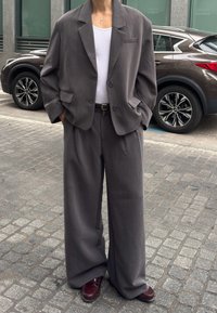 Persoană îmbrăcată într-un costum gri supradimensionat cu pantaloni largi, maiou alb, curea neagră și pantofi maro, stând pe o stradă pavată cu o mașină parcată în spate.