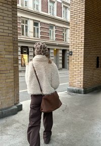 Person iført leopardmønstret hat, hvid fluffy jakke, brune bukser og bærende en brun taske går gennem en bue ved siden af en murstensbygning.