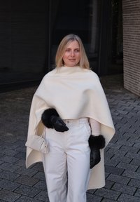 Femme blonde portant une cape blanche, un pantalon blanc, des gants noirs avec des poignets en fourrure, tenant un petit sac à main blanc, debout sur un pavé en pierre à l'extérieur.