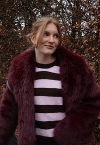 Jeune femme aux cheveux clairs portant un manteau en fausse fourrure violet par-dessus un pull rayé noir et rose, debout à l'extérieur près de buissons secs et feuillus.