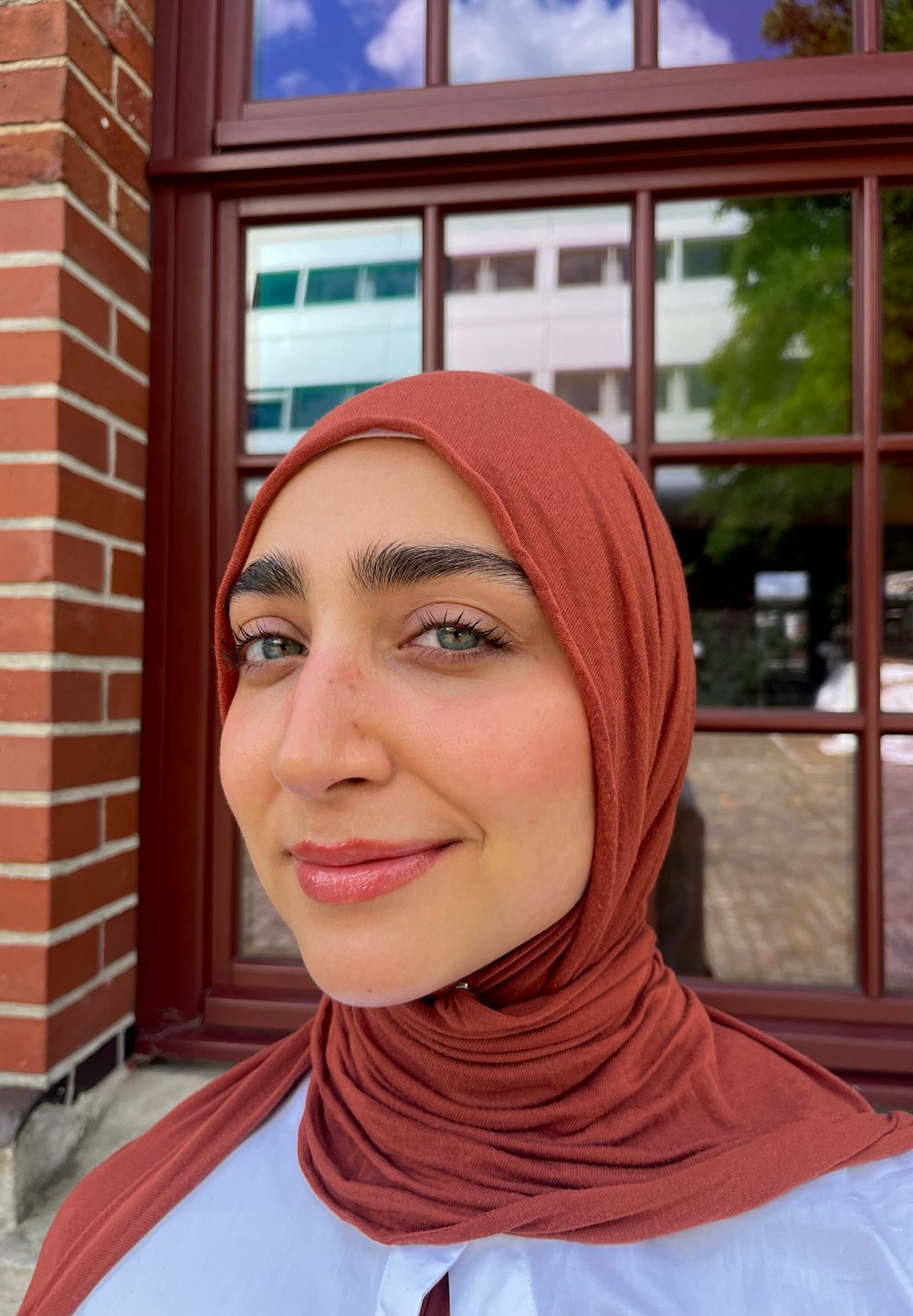 Vrouw met een roestkleurige, gestructureerde hijab en een lichtblauw topje, met een egale huid, opvallende wenkbrauwen en groene ogen, tegen een bakstenen muur.