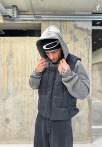 Gilet imbottito nero con colletto alto, indossato sopra una felpa grigia. Il modello indossa un berretto nero e si trova in un ambiente in cemento testurizzato.