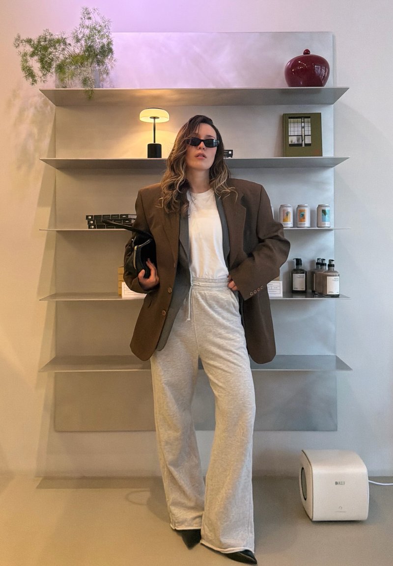 Γυναίκα που φοράει υπερμεγέθη καφέ σακάκι, λευκό t-shirt, γκρι φόρμα και γυαλιά ηλίου, στέκεται μπροστά σε ράφια τοίχου με διακοσμητικά και μπουκάλια.