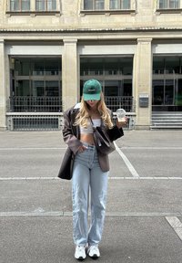 Μοντέλο φορώντας ένα καφέ καρό υπερμεγέθες σακάκι, λευκή κοντή μπλούζα, ανοιχτό μπλε τζιν και πράσινο καπέλο, κρατώντας ένα ποτό, με λευκά αθλητικά παπούτσια.