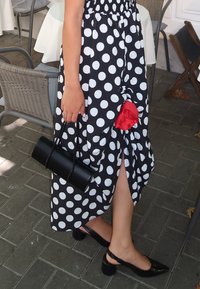 Donna in un abito a pois bianchi e neri, con una pochette nera, indossando scarpe nere con tacco basso, con un dettaglio in tessuto rosso sulla fessura del vestito.