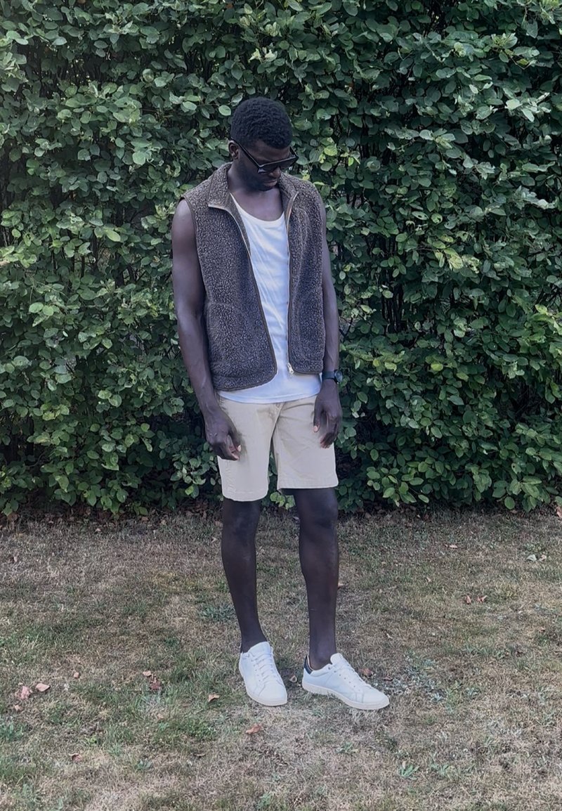 Homme portant des lunettes de soleil, un gilet marron sans manches, une chemise blanche, un short beige et des baskets blanches, debout sur l’herbe devant une haie verte et feuillue.