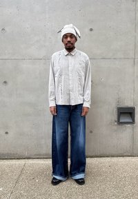 Homme debout contre un mur en béton, portant une chemise claire à motifs, un jean bleu ample, des chaussures noires et un turban assorti en tissu léger.