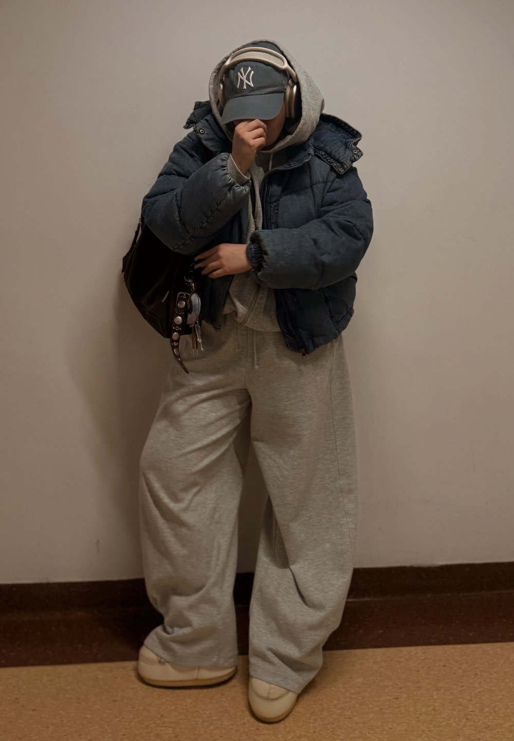 Geacă puffer albastru închis, hanorac gri, pantaloni de trening gri deschis, pantofi slip-on albi, șapcă gri cu logo, geantă mare neagră, căști.