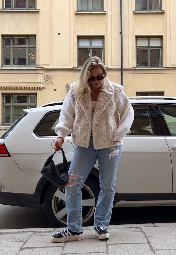 Faux fur creme overtøj med en dobbeltknap-design, parret med lyseblå slidte jeans og sorte sneakers. Holder en lille sort taske.
