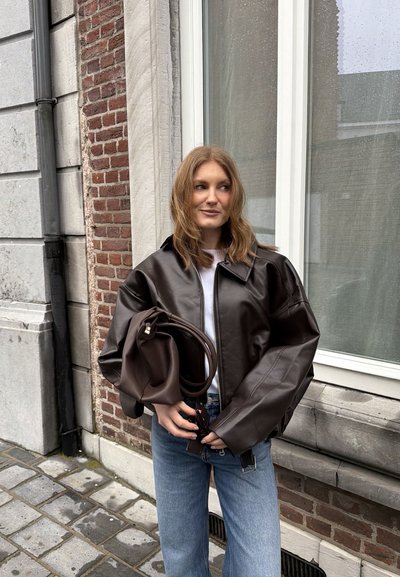 Mujer con cabello castaño claro que lleva una chaqueta de cuero marrón oscuro extragrande y pantalones vaqueros azules, sosteniendo un bolso de cuero marrón oscuro a juego.