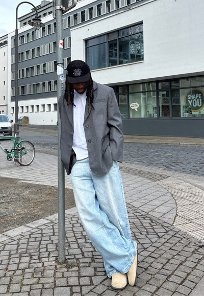 Blazer gris sobre una camisa blanca, combinado con jeans de pierna ancha de color azul claro y zapatos beige. Gorra negra con logo. Entorno urbano con adoquines.