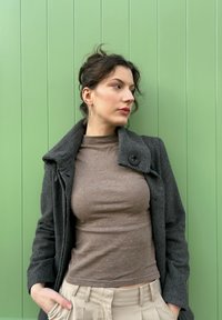 Manteau en laine gris foncé avec un grand bouton, haut à col roulé marron, et pantalon plissé beige, sur fond de panneau vertical vert.