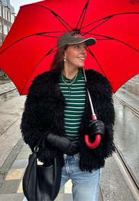 Palton de blană neagră, cămașă cu dungi verzi și blugi. Ține în mână o umbrelă roșie cu un mâner curb, purtând mănuși negre și o șapcă.