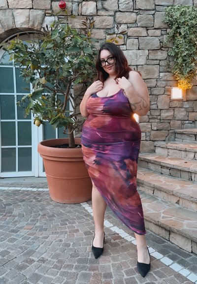 Vestido midi tie-dye en tonos púrpura y rosa, sin mangas con un diseño drapeado. La modelo lo combinó con tacones puntiagudos negros, posando junto a una pared de piedra.