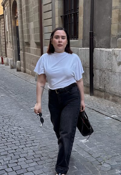 Top blanco de manga corta, vaqueros oscuros de cintura alta y bolso negro. Caminando por una calle de piedra con gafas de sol en la mano.