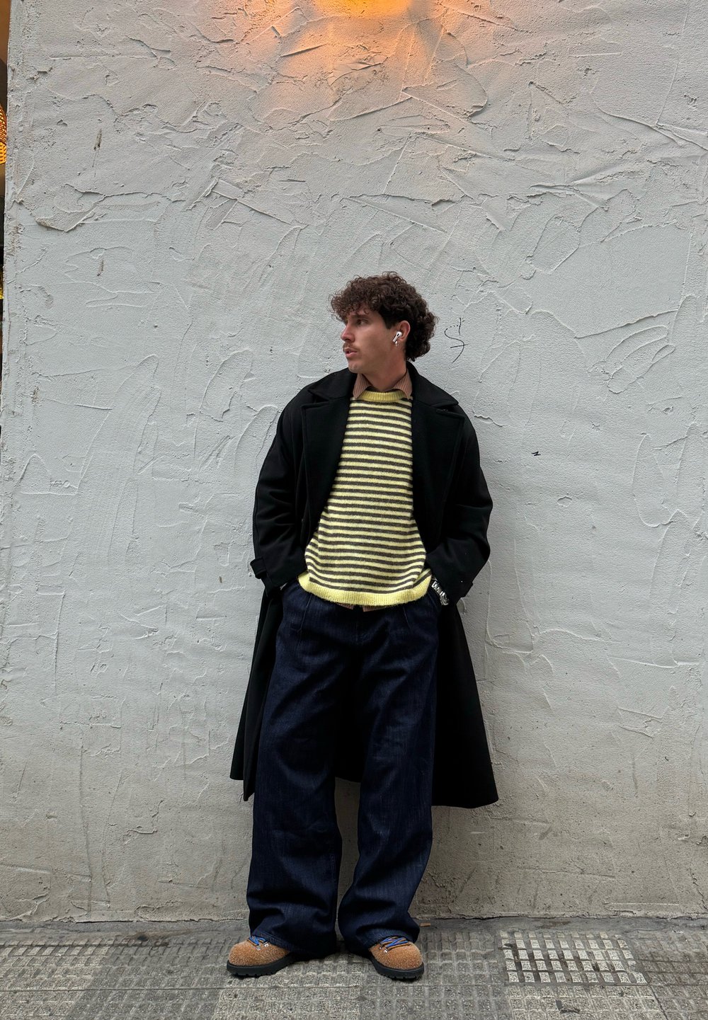 Jeune homme aux cheveux bouclés portant un pull jaune à rayures, un manteau noir, un pantalon large noir et des chaussures marron, debout devant un mur texturé.
