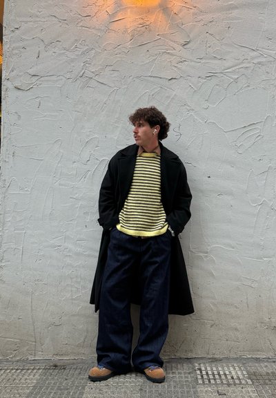 Joven con cabello rizado que lleva un suéter amarillo a rayas, un abrigo negro, pantalones anchos negros y zapatos marrones, de pie frente a una pared texturizada.