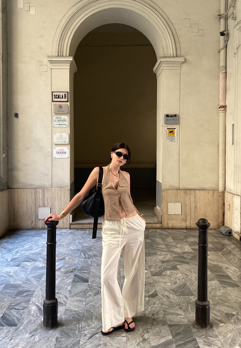 Μοντέλο φορώντας ένα μπεζ κοντό τοπ και λευκά παντελόνια σε φαρδιά γραμμή, με μαύρα γυαλιά ηλίου και μια μεγάλη μαύρη τσάντα, στέκεται δίπλα σε μια μαύρη κολώνα σε ένα διάδρομο.