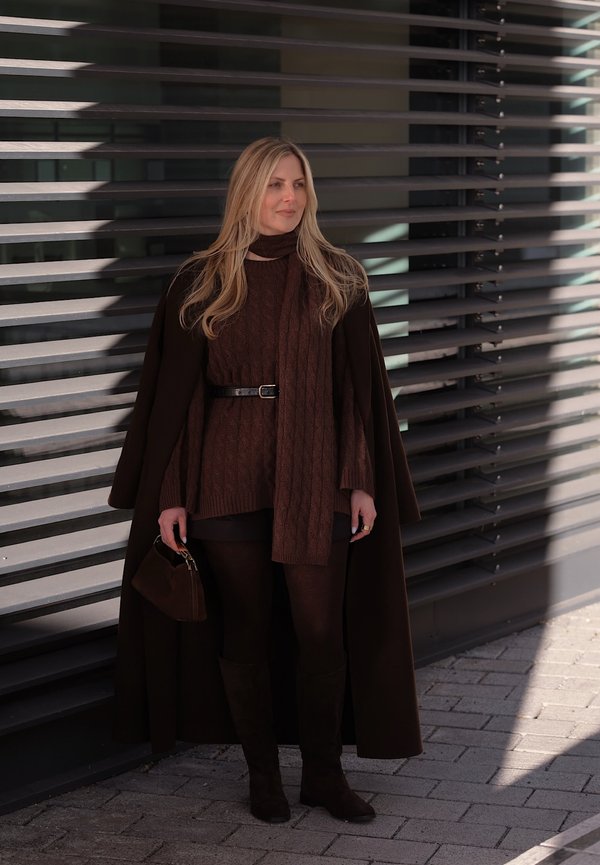 Donna con lunghi capelli biondi in piedi accanto a una parete moderna in lamelle di metallo, indossa un cappotto marrone scuro, un vestito a maglia, una sciarpa, calze e stivali, tiene in mano una piccola borsa.