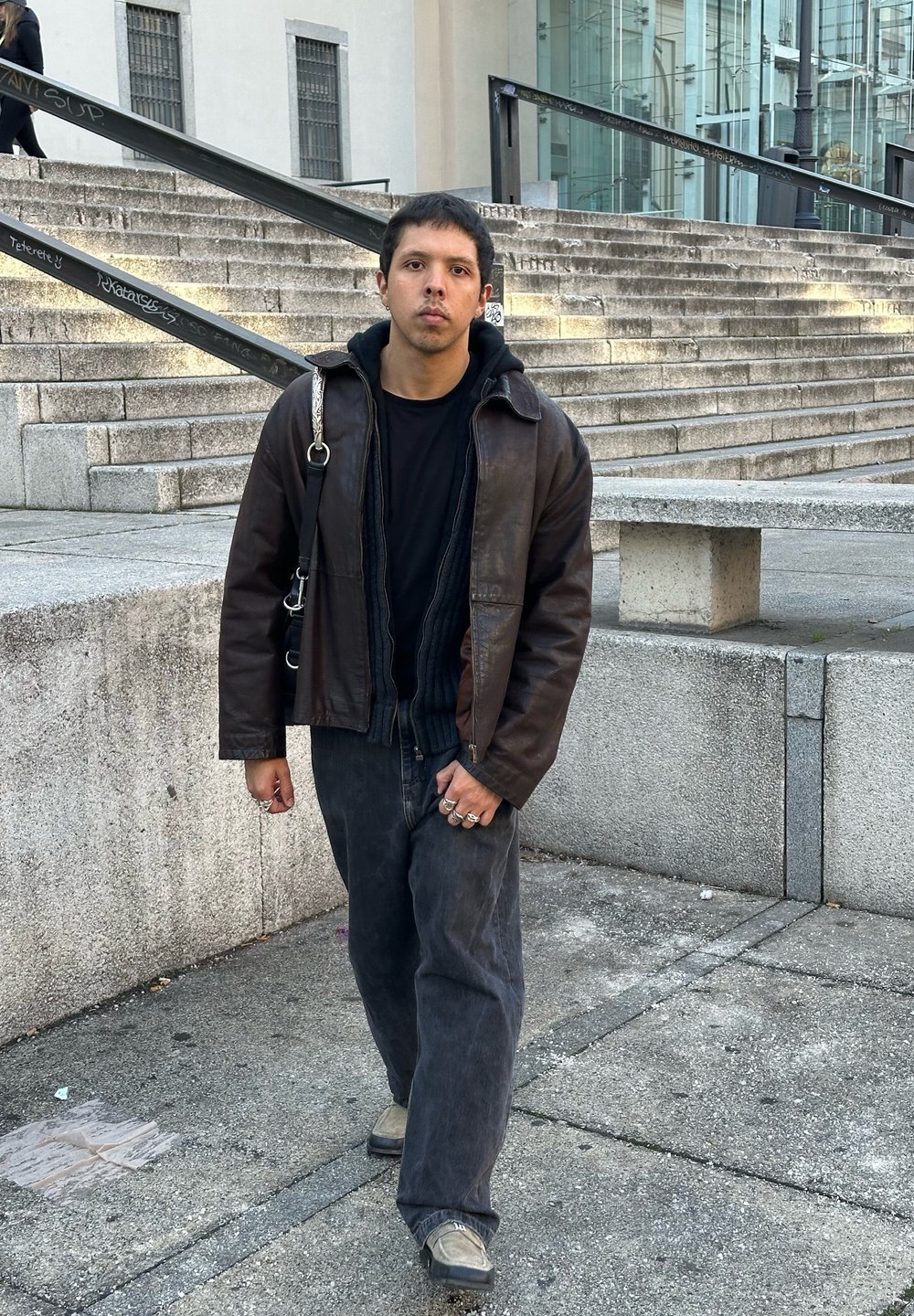 Brown leather jacket worn over a black sweater, teamed with loose-fitting black trousers and grey shoes. Distinctive rings adorning the fingers, set against a textured backdrop.
