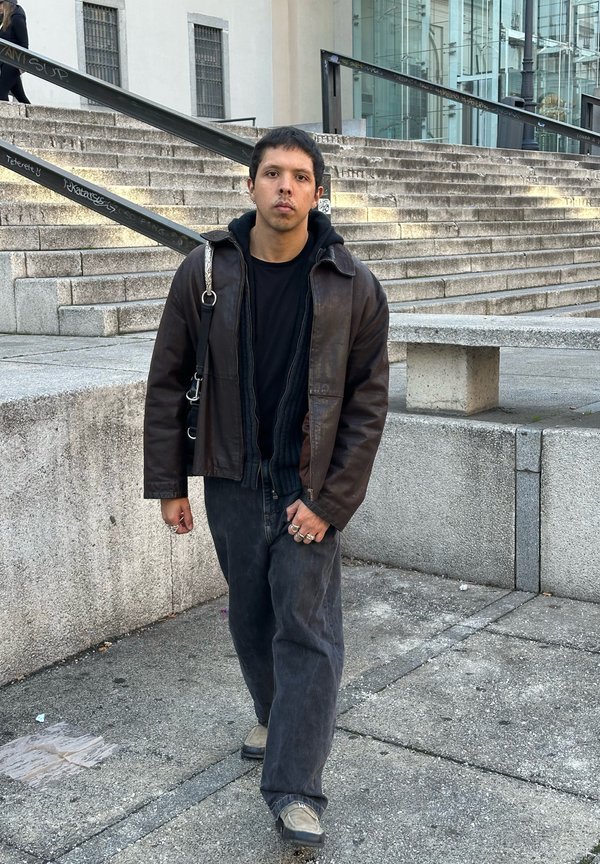 Brown leather jacket worn over a black sweater, teamed with loose-fitting black trousers and grey shoes. Distinctive rings adorning the fingers, set against a textured backdrop.