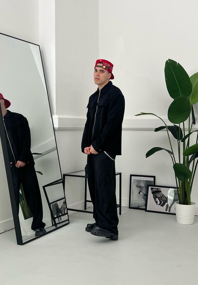 Modelo con una chaqueta negra con cremallera y pantalones sueltos. Gorra de béisbol roja con logo. Zapatos de plataforma negros. Reflejo en un gran espejo. Planta verde cercana.