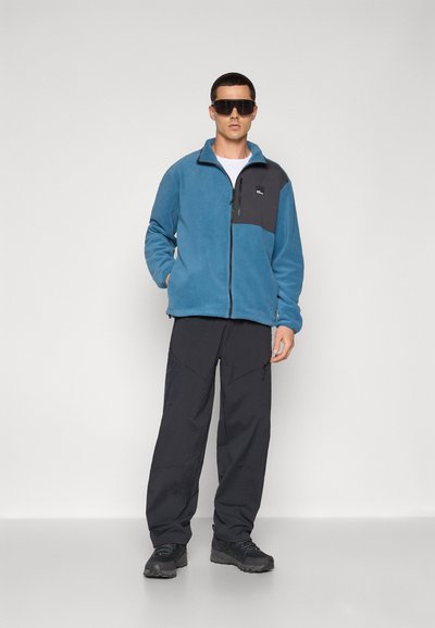 Chaqueta de forro polar azul con un panel negro, cierre de cremallera y bolsillo de parche, combinada con pantalones cargo oscuros y zapatos de senderismo negros.