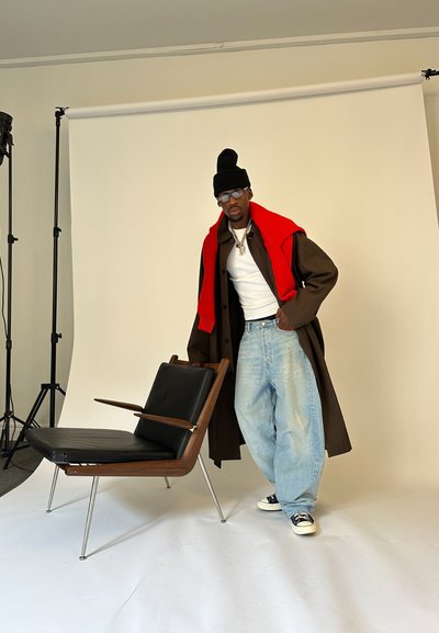 Hombre con gorro negro, gafas, abrigo marrón, suéter rojo sobre los hombros, camisa blanca, jeans holgados y zapatillas de deporte, de pie al lado de una silla de estilo moderno de mediados de siglo.