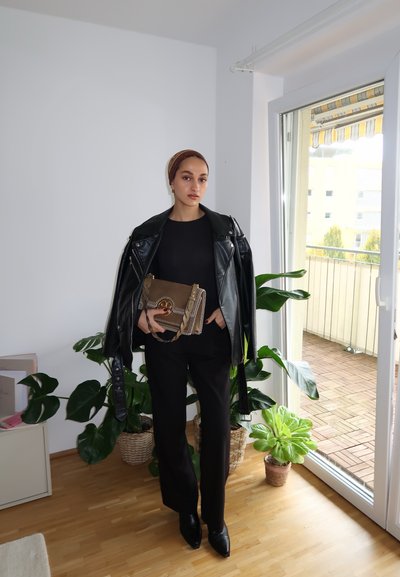 Chaqueta de cuero negra sobre una blusa negra, combinada con pantalones negros a medida. Sostenido un bolso beige con detalles de cadena. Entorno interior con plantas.