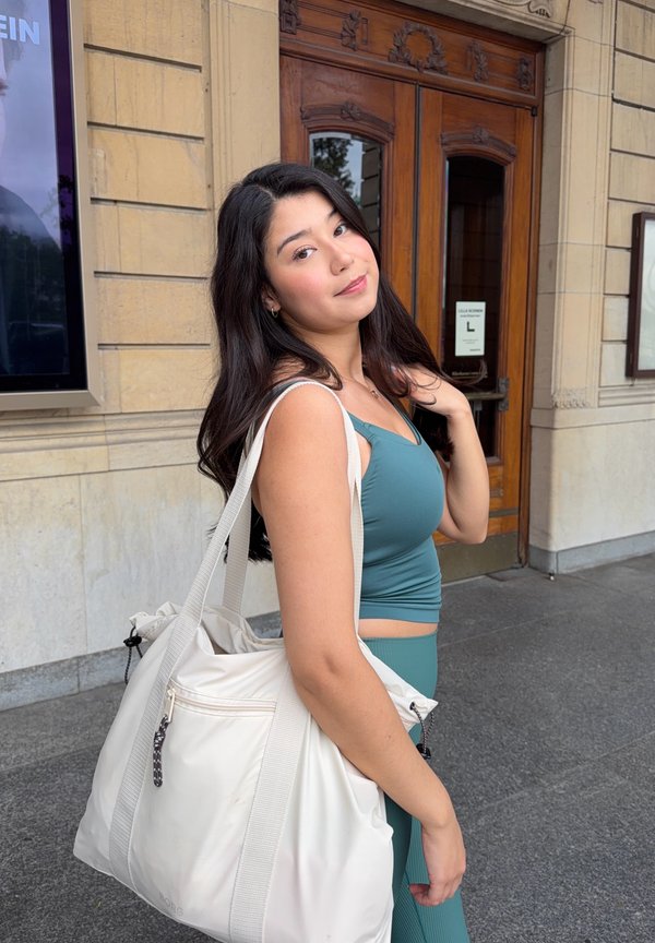 Une femme vêtue d’une tenue turquoise pose devant un bâtiment, portant un grand sac blanc à l’épaule.