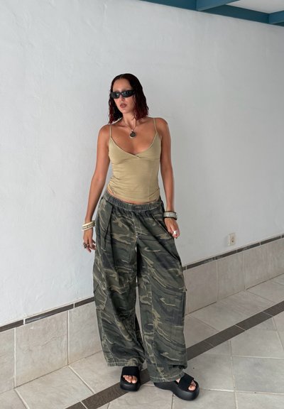 Top sin mangas ajustado, pantalones de camuflaje de corte holgado, sandalias negras de plataforma con suelas gruesas y pulseras en capas. Fondo neutro.