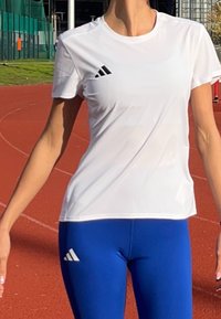 Idrettsutøver iført hvit Adidas-skjorte og blå Adidas-tights som står på en rød utendørs løpebane.