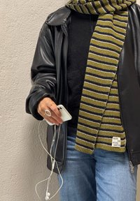 Person med svart skinnjakke, stripete grønn og svart skjerf, og jeans som holder en hvit smarttelefon med ørepropper plugget inn.