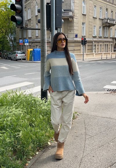 Mujer con un suéter a rayas azul y blanco, pantalones beige y botas, camina por una acera de la ciudad cerca de un semáforo y un paso de peatones.