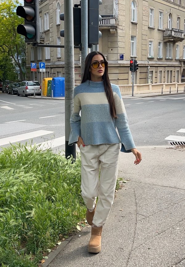 Kvinde iført en blå- og hvidstribet sweater, beige bukser og støvler går på en byfortov nær et trafiklys og fodgængerfelt.