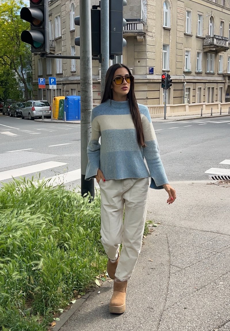 Frau trägt einen blau-weiß gestreiften Pullover, beigen Hosen und Stiefel und geht auf einem Gehweg in der Stadt in der Nähe einer Ampel und eines Fussgängerstreifens.