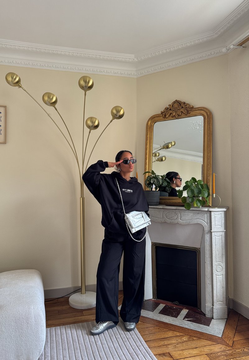 Personne portant une tenue noire, des lunettes de soleil et un sac argenté, debout près d'une cheminée blanche ornée avec un miroir encadré d'or et une grande lampe sur pied en laiton.