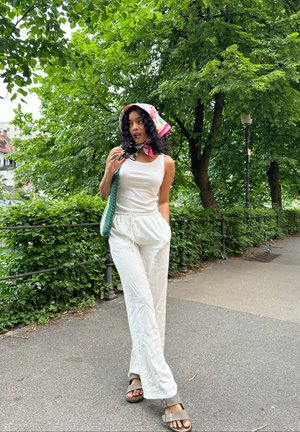 Top blanco sin mangas y pantalones blancos holgados, complementados con un pañuelo de cabeza colorido y un bolso tejido verde. Sandalias marrones con detalle de tiras.