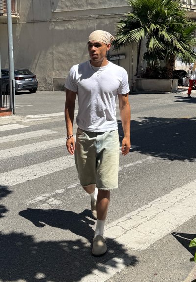 Camiseta blanca y pantalones cortos verdes desgastados, calcetines blancos y zuecos beige. Bandana con un patrón. Fondo de calle y palmera.