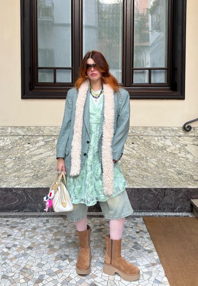 Blazer gris de gran tamaño con cuello de piel sintética, vestido floral verde menta, pantalones cortos de denim, botas de ante marrón hasta la rodilla y un bolso blanco adornado con llaveros.