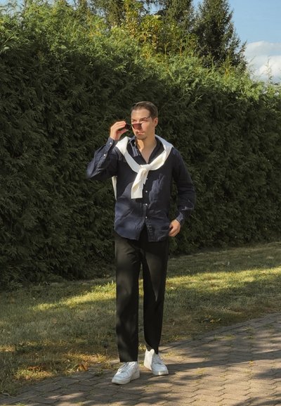 Hombre vistiendo una camisa de botón azul oscuro, un suéter blanco colgado sobre los hombros, pantalones negros y zapatillas blancas, posando al aire libre.